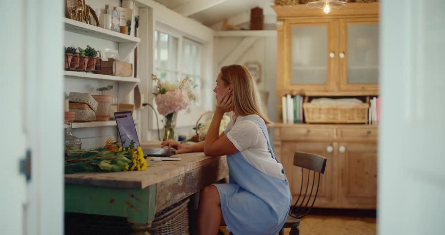 A female florist works on her laptop computer, managing her small business from her charming rustic flower workshop