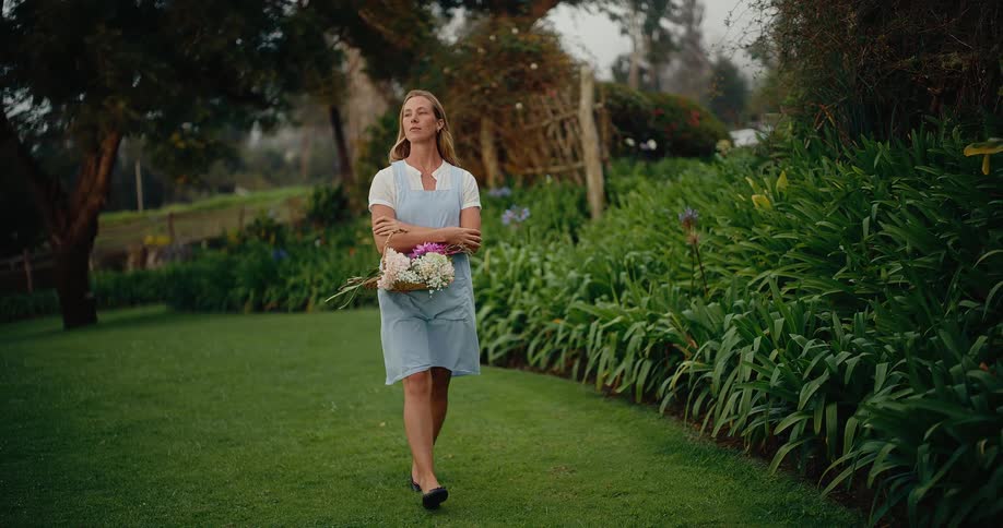 A graceful woman in a blue apron walks in slow motion through a lush green garden carrying a basket of freshly picked flowers