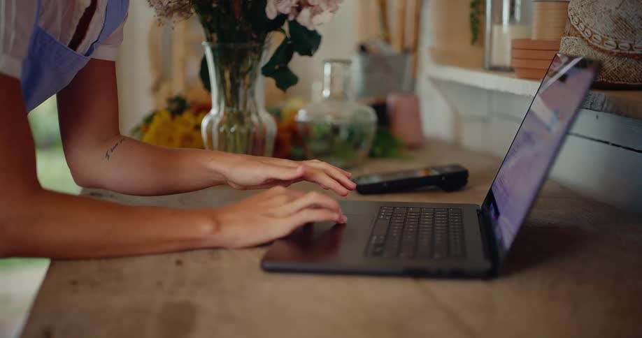 Close up of a florist's hands working on a laptop at a rustic wooden desk before arranging a bouquet of flowers in a vase