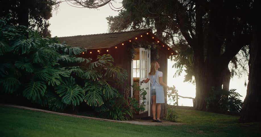 A woman wearing an apron and holding flowers walks into a small, quaint cottage in a garden at dusk