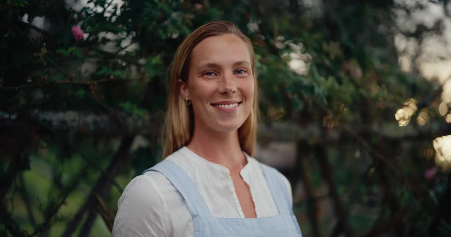 A beautiful young woman in a blue apron smiles peacefully in her lush garden at dusk