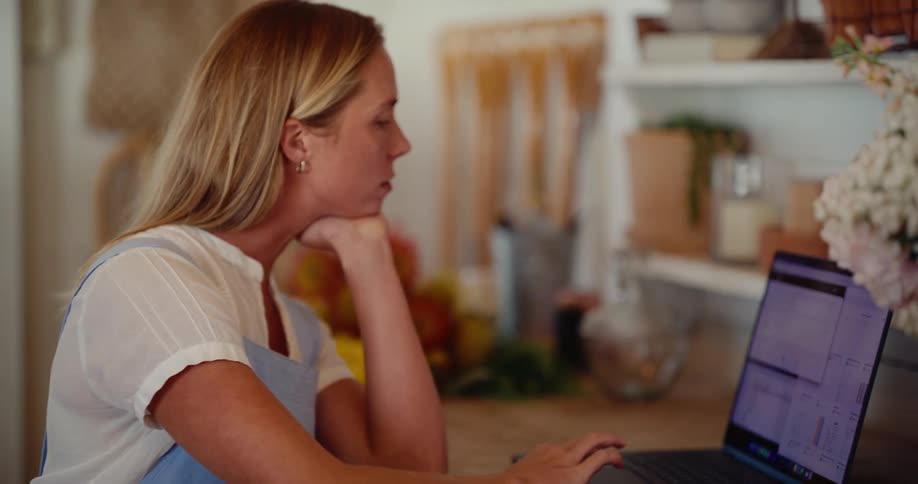 A female florist wearing an apron works on her laptop and uses a payment terminal to manage her business finances in her flower shop
