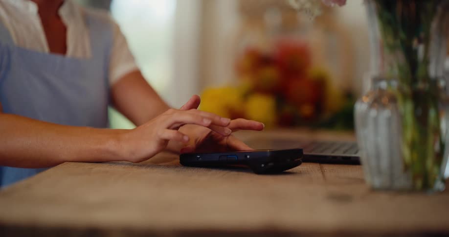 Close up of a smiling female florist in her shop, tilting down to her hands as she uses a POS terminal and laptop to manage her business