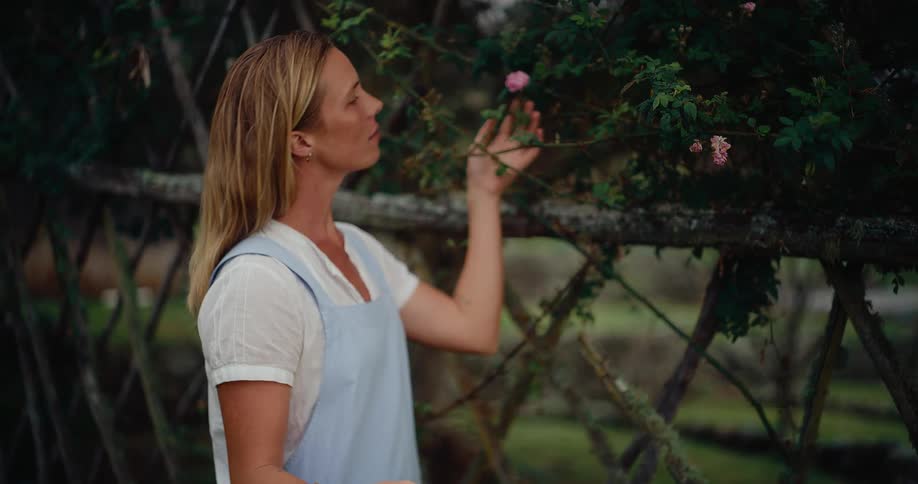 A serene woman in a blue apron holding a basket of fresh flowers gently smells a pink rose in her garden