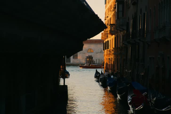 Moored gondolas line a tranquil Venetian canal during a beautiful golden hour sunset as a boat passes in the background