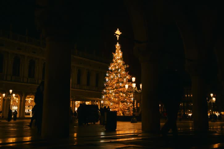 Silhouetted figures walk through a historic European square illuminated by a grand Christmas tree at night