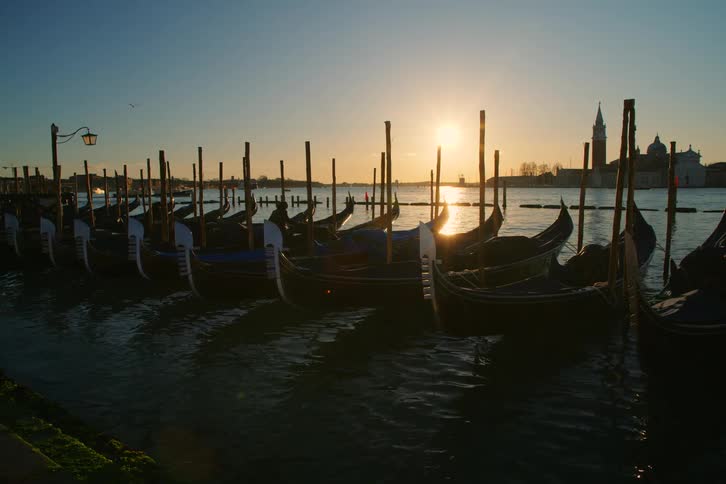 Gondolas moored in a row gently rock on the water in Venice, silhouetted by the setting sun