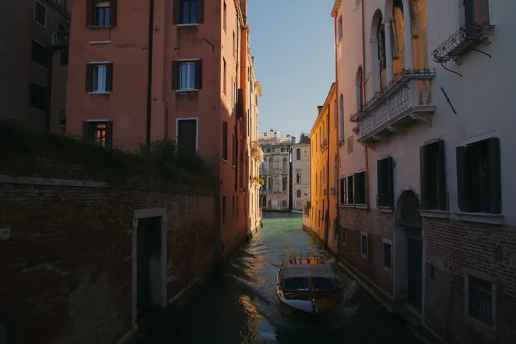 A classic wooden water taxi navigates a narrow, sun-drenched canal between historic buildings in Venice, Italy
