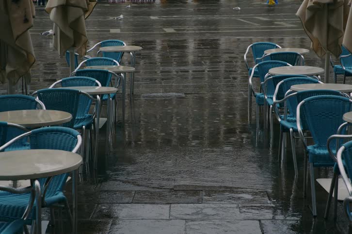 Rain falls on the wet stone ground of an empty outdoor cafe with blue chairs and closed umbrellas