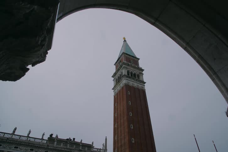 A low-angle view of the iconic St. Mark's Campanile bell tower in Venice, framed by a stone archway under a grey, overcast sky