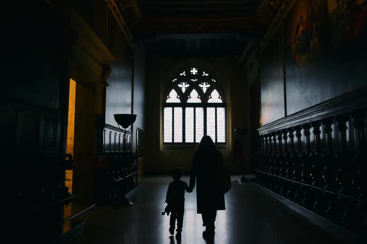 A silhouetted mother and child hold hands as they slowly walk down a dark, historic hallway towards a bright, arched window