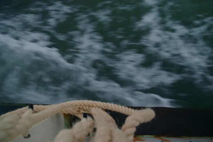A point-of-view shot from the side of a boat captures the fast-moving wake churning in the ocean with a coiled rope in the foreground
