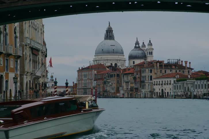 A classic water taxi navigates Venice's Grand Canal past the Basilica di Santa Maria della Salute, viewed from beneath a bridge on an overcast day