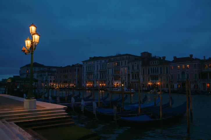 Gondolas are moored in a Venice canal as evening falls and an ornate street lamp casts a warm glow