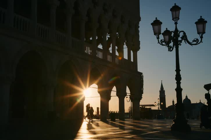 The rising sun casts a brilliant flare through the arches of the Doge's Palace in a nearly empty St. Mark's Square in Venice, Italy