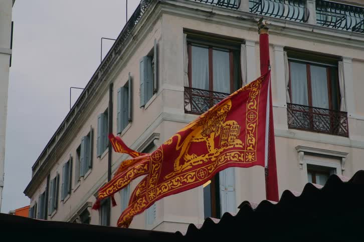 The historic flag of Venice featuring the Lion of Saint Mark waves in slow motion against a classic building on an overcast day