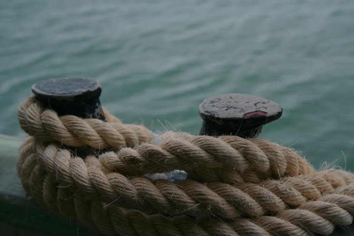 A close-up of a thick nautical rope wrapped around two metal mooring bollards on a boat, with the sea moving in the background