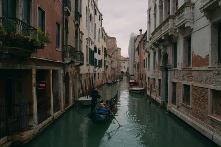A gondolier wearing a traditional hat rows a boat in slow motion down a picturesque canal between old Italian buildings in Venice