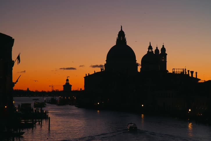 A silhouette of the Basilica di Santa Maria della Salute against a vibrant orange sunset sky as a water taxi travels along the Grand Canal in Venice, Italy