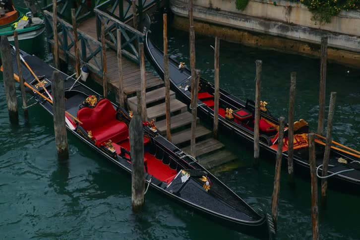 Two empty gondolas with plush red seats gently sway while docked at a wooden pier in a Venice canal