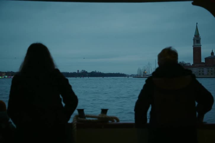 A silhouetted couple watches the Venice skyline pass by from a boat on a moody, overcast evening