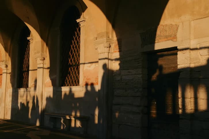 The golden light of sunset casts the moving shadows of a passing crowd across the ancient arched walls of a historic building