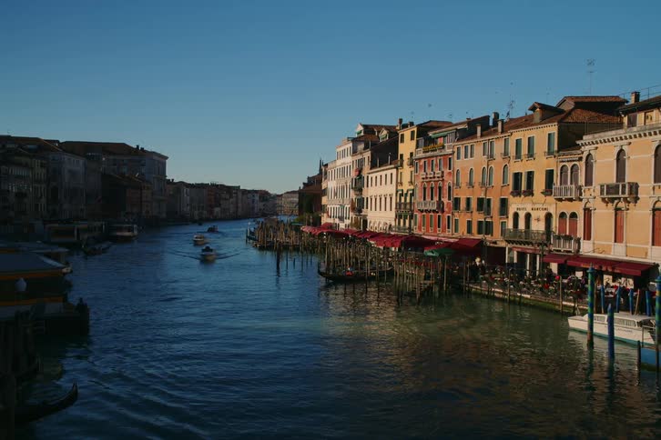 Boats and gondolas traverse the iconic Grand Canal in Venice, Italy, flanked by sunlit historic buildings under a clear blue sky