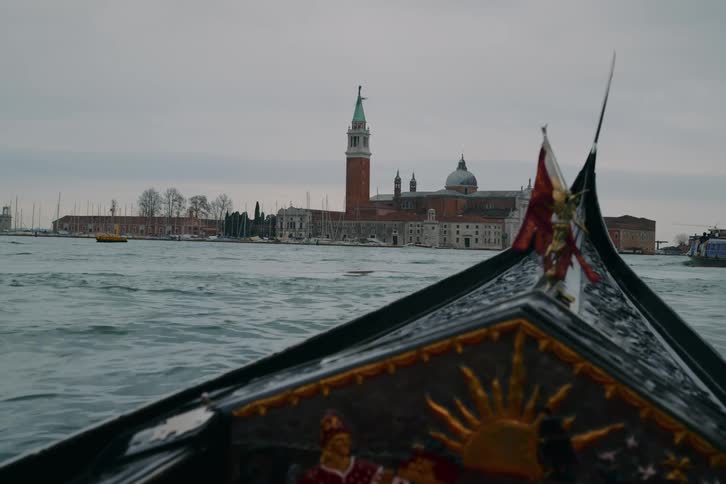 A Venetian gondola gently rocks on the choppy lagoon, offering a classic point-of-view of the San Giorgio Maggiore island on an overcast day