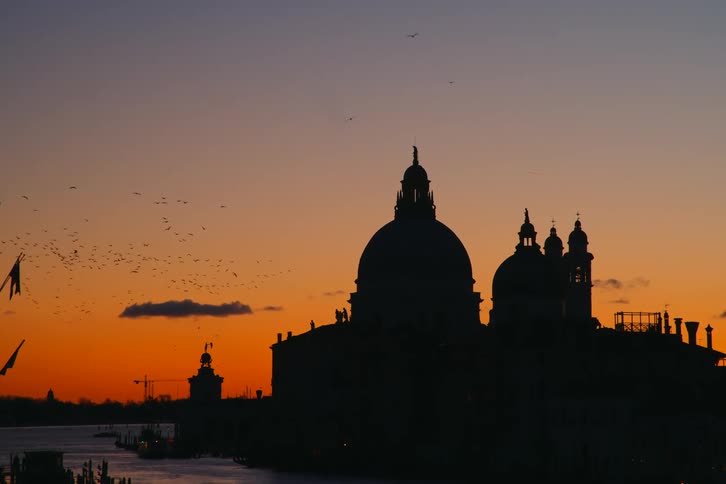 The silhouette of the Basilica di Santa Maria della Salute in Venice is framed by a vibrant orange sunset as a flock of birds flies by