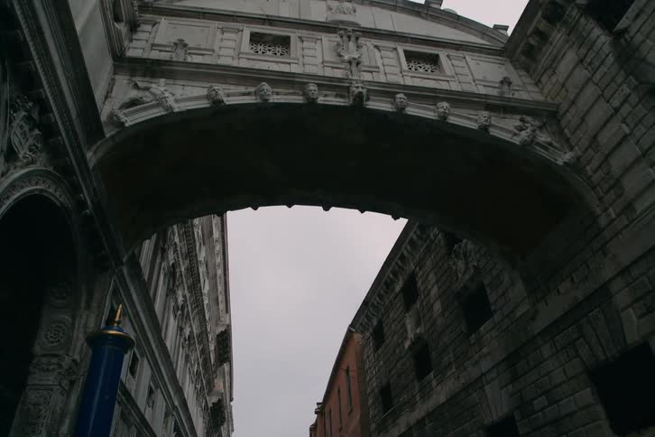 A slow, gliding shot from a low angle passes underneath the iconic Bridge of Sighs in Venice, Italy on an overcast day