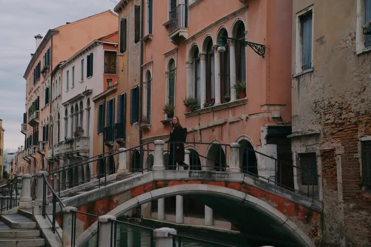A woman leisurely crosses a picturesque bridge spanning a quiet canal amidst the historic, colorful buildings of Venice