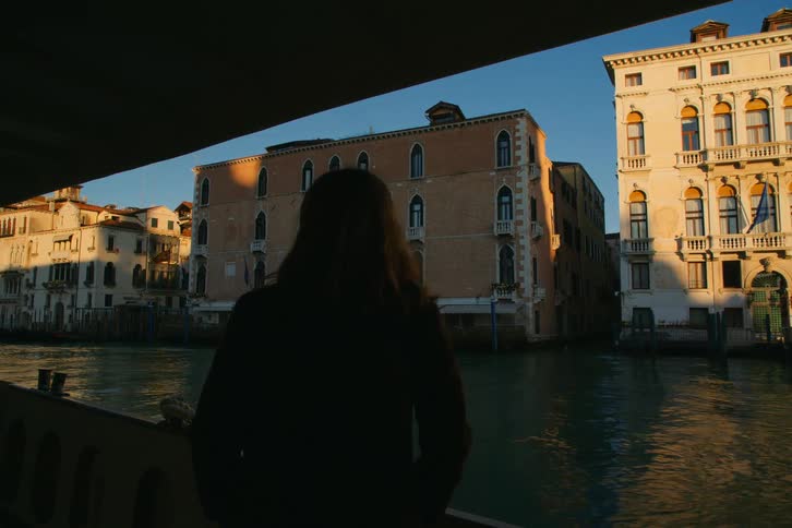 A silhouetted woman watches the historic buildings of Venice pass by from a boat during a golden sunset