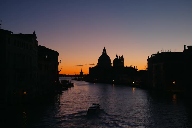 A lone water taxi cruises down the Grand Canal in Venice, Italy at dusk, with the Basilica di Santa Maria della Salute silhouetted against the vibrant sunset