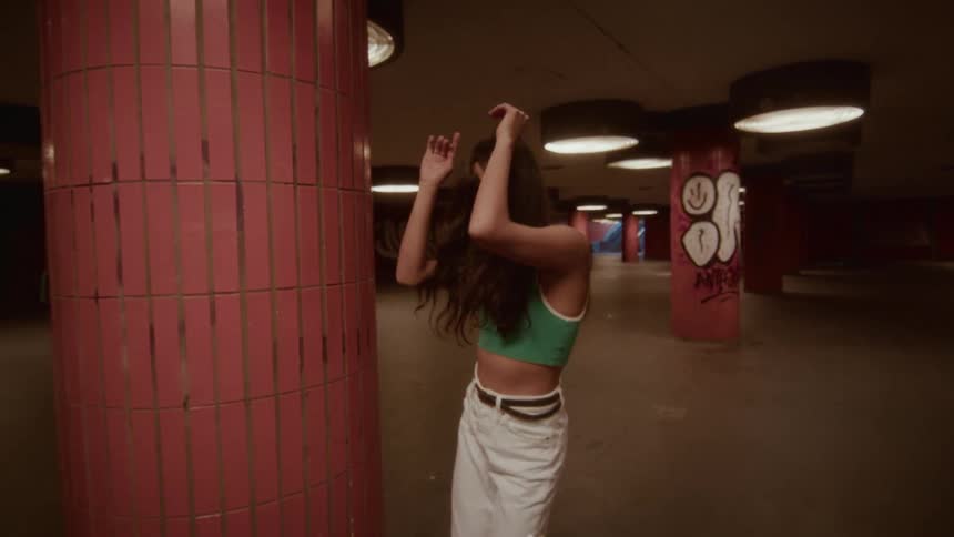 A carefree woman in retro roller skates dances and grooves next to a tiled pillar in a dimly lit, urban underpass