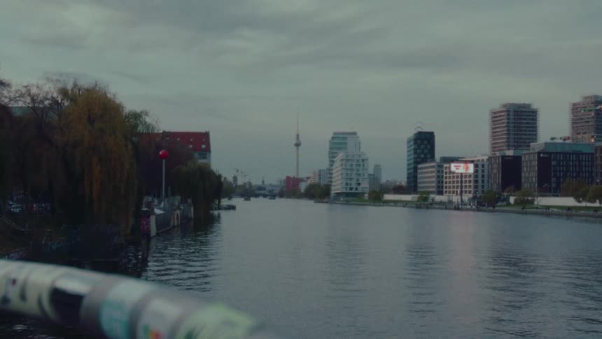 A moody, static shot of the Spree river in Berlin, with the city's skyline and the Fernsehturm visible in the distance on a cloudy day