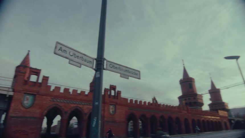 Pedestrians walk along the historic Oberbaum Bridge in Berlin, with the camera tilting up to a street sign in vintage-style footage