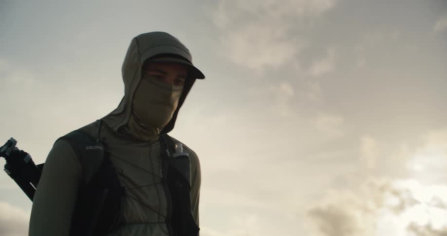 A mysterious hiker in a balaclava stands pensively against a bright, cloudy sky in slow motion