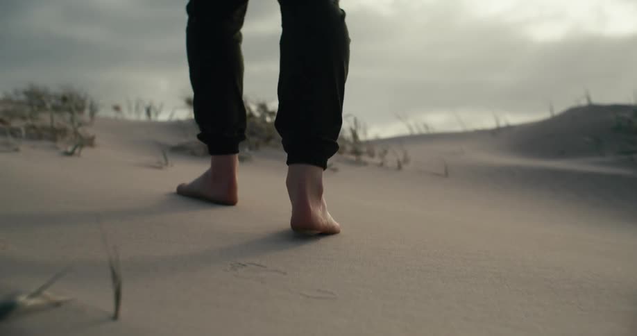 A low-angle slow-motion view follows a person's bare feet walking across a sandy dune under a cloudy sky