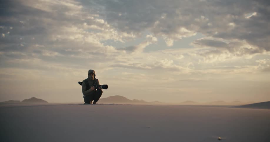 A dedicated photographer kneels in a vast, windy desert, waiting patiently for the perfect shot at dusk