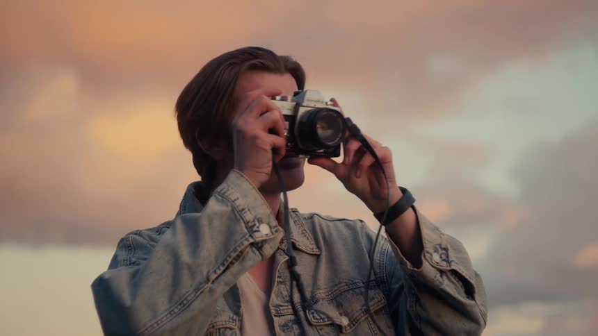 A young man in a denim jacket smiles and takes a photo with a vintage film camera against a beautiful sunset sky