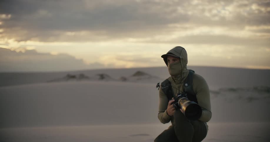 A dedicated photographer with a telephoto lens kneels in vast sand dunes, scanning the horizon at sunset