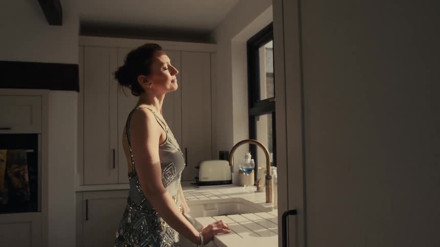 An elegantly dressed woman takes a quiet moment to feel the warm sunlight on her face while standing in her modern kitchen