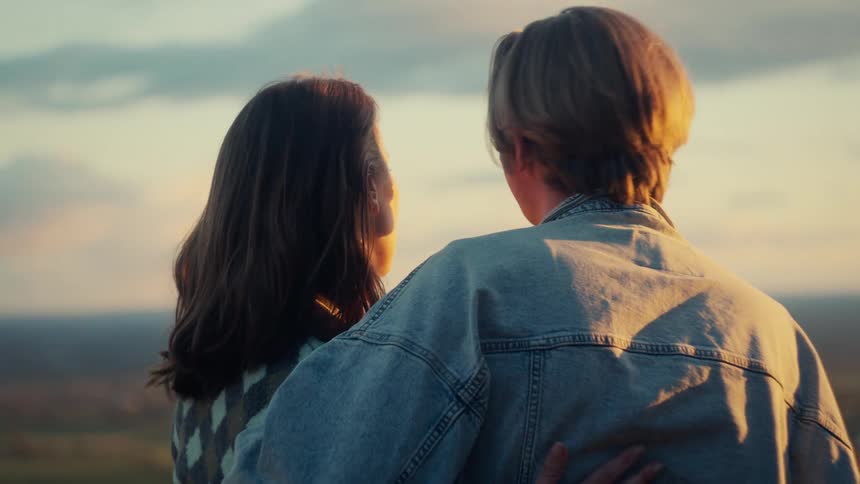 A slow-motion rack focus reveals a romantic young couple embracing as they watch the beautiful golden hour sunset over the countryside