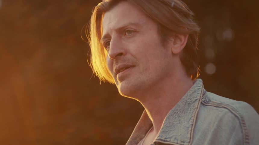 A pensive man with long hair smokes a cigarette in slow motion, backlit by a warm golden hour sunset
