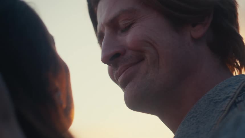 A man gazes lovingly at his partner as her hand gently caresses his face during a warm sunset