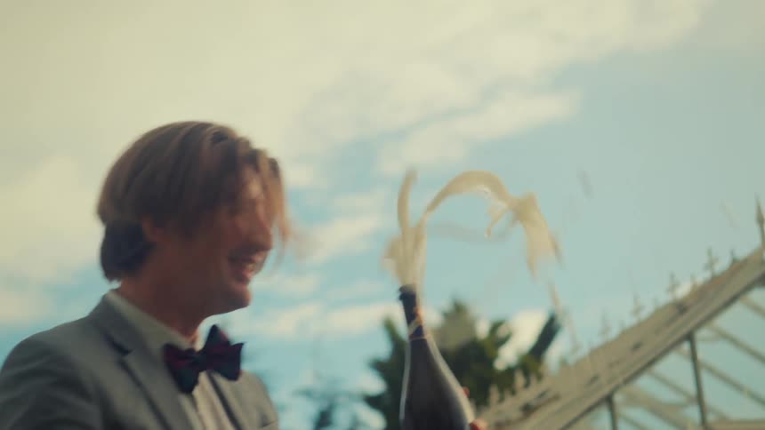 A jubilant man in a suit and bow tie pops a champagne bottle in slow motion, celebrating with a spray of foam