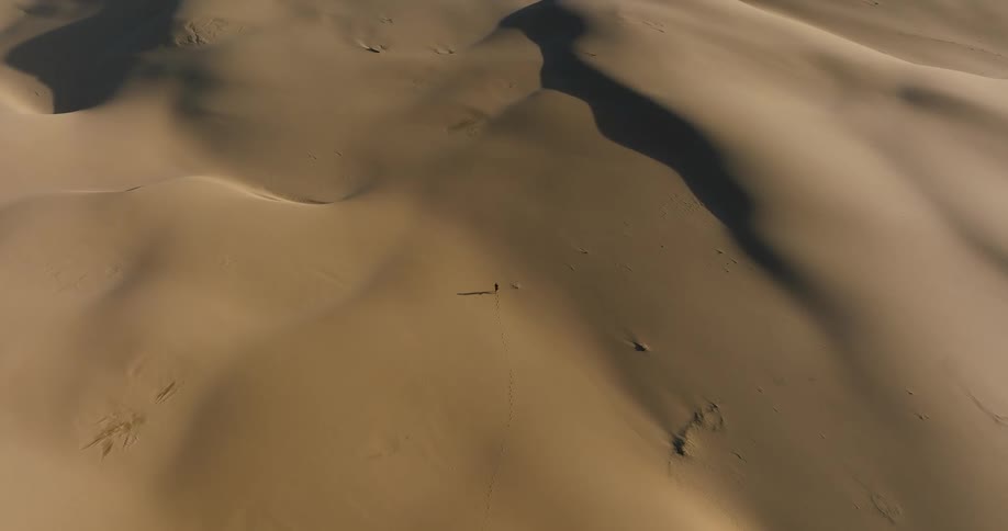 A high-angle static aerial shot shows a single person dwarfed by the immense and beautiful sand dunes as they walk through the vast desert
