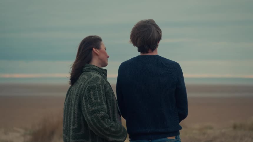A young couple shares a quiet, intimate moment while standing on a windy beach and looking out at the ocean