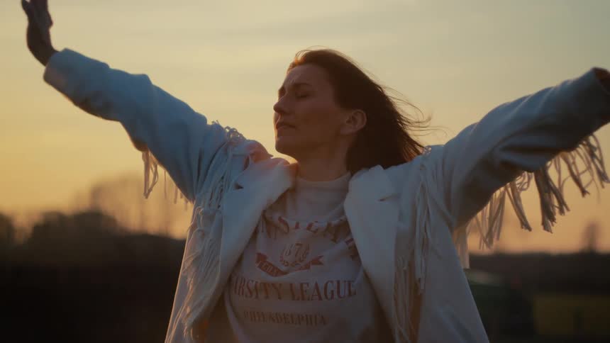 A free-spirited woman in a fringed jacket embraces the golden hour sunset with her arms outstretched in slow motion