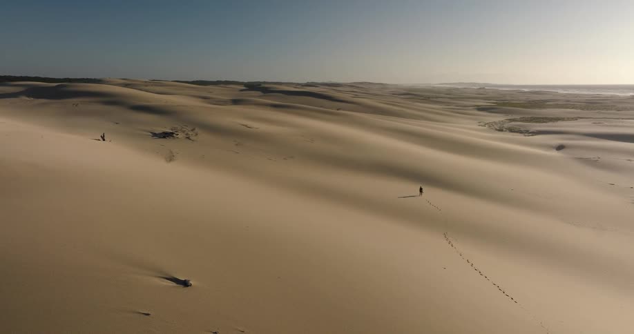A lone hiker treks through endless golden sand dunes under a clear sky